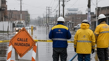 Tres operarios de Luz del Sur con uniformes y cascos parados frente a un cartel naranja de "CALLE INUNDADA" y cinta de "PELIGRO" en una vía anegada con postes de luz