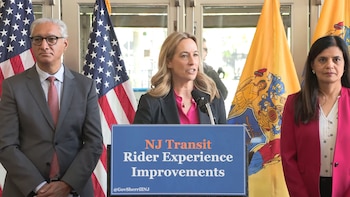 Mikie Sherrill en un podio con un micrófono, flanqueada por dos personas. Detrás, banderas de EE. UU. y Nueva Jersey. El podio dice "NJ Transit Rider Experience Improvements"