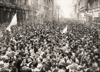 Fotografía en blanco y negro de una multitud de personas que camina por una calle flanqueada por edificios con algunas banderas