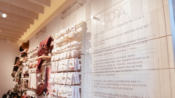 Interior de una tienda de accesorios con estantes blancos llenos de anillos, pendientes, collares, bolsos y sombreros; un gran panel de vidrio con texto es visible a la derecha