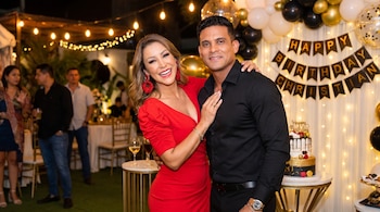 Karla Tarazona de rojo y Christian Domínguez de negro, posan en fiesta de cumpleaños con cartel, globos, luces y pastel.