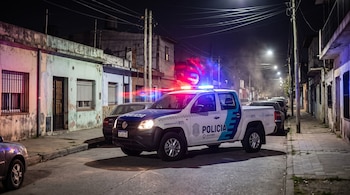 Camioneta policial con luces azules y rojas encendidas en una calle asfaltada de noche, entre casas bajas y postes de tendido eléctrico.