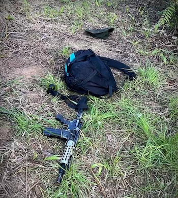 Un fusil de asalto con cargador, una mochila negra con detalles azules y una gorra verde oscuro tirados en el suelo cubierto de hierba y tierra seca