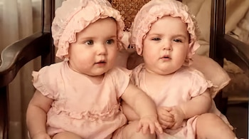 Dos bebés con vestidos y gorros rosados están sentadas juntas sobre una silla de madera con respaldo de rejilla.