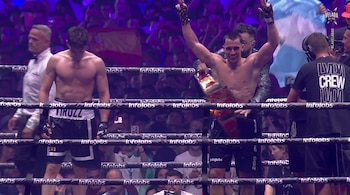 Dos hombres en un ring de boxeo con cuerdas. Uno sonríe con brazos en alto, cinturón y bandera celeste y blanca. El otro está de pie, cabizbajo. Multitud.