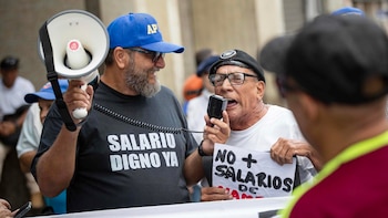 Dos hombres protestando: uno con gorra azul y camiseta 'SALARIO DIGNO YA' sostiene un megáfono, el otro con gorra negra habla a un micrófono y sujeta un cartel