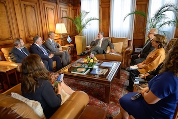 Grupo de ocho personas sentadas en sofás de cuero. En el centro, una mesa de café de madera y vidrio con libros y flores. Paredes con paneles de madera