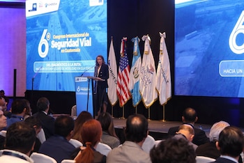 Un expositor junto a un atril y dos pantallas grandes presenta información ante un público sentado en un salón de conferencias