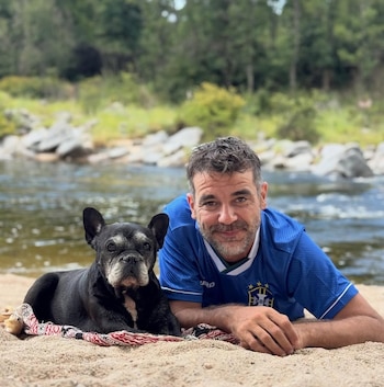 Stonnie Dennis, con camiseta azul, yace en la arena junto a un bulldog francés negro con hocico gris a orillas de un río