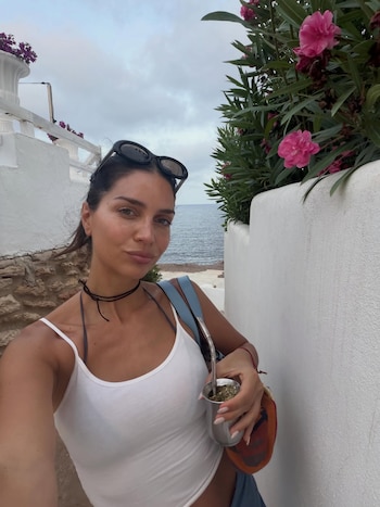 Mujer morena con gafas de sol en la cabeza, camiseta de tirantes blanca, choker negro, sostiene un mate y una bombilla. Hay vegetación con flores rosas y el mar