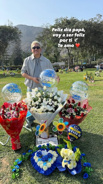 Hombre rubio con gafas de sol, sosteniendo un globo transparente, de pie junto a tres ramos de flores, varios globos y una lápida en un cementerio