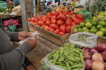 Unas manos cuentan billetes y monedas junto a tomates, limones, cebollas y arvejas expuestos en un puesto de madera.