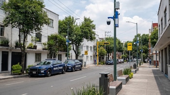 Calle urbana con edificios blancos, árboles verdes, dos patrullas azules de la policía, cámaras de videovigilancia en postes y mobiliario público moderno en Venustiano Carranza.