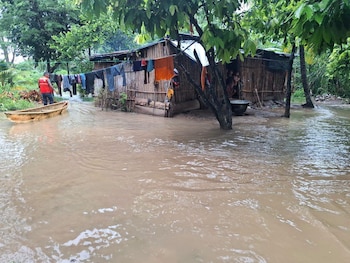 Una casa de madera y bambú parcialmente sumergida en agua marrón turbia. Ropa colgada afuera. Un hombre en bote en el agua y niños en la entrada de la casa