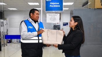 Hombre de ATU con chaleco azul y blanco entrega un bolso de mano color crema a una mujer sonriente, bajo un letrero que dice 'Objetos Perdidos'