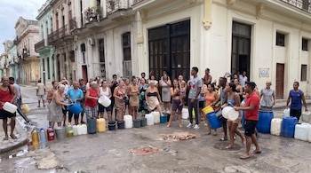Personas reunidas en una calle junto a alineaciones de bidones, recipientes de plástico y cubos vacíos sobre el asfalto.