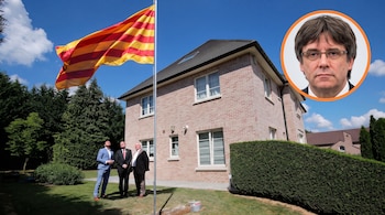 Tres personas miran una bandera catalana izada frente a una casa de ladrillo, con un retrato circular de Carles Puigdemont arriba a la derecha.