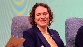 Liz Reid, con cabello rizado y una chaqueta azul marino sobre una camiseta beige, sonríe ampliamente sentada en una silla gris. El logo de Google es visible