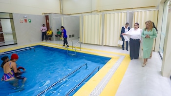 Piscina interior con barandales y borde amarillo. Dos personas en el agua, una con gorro rojo. Seis adultos caminan o esperan en el área alrededor de la piscina