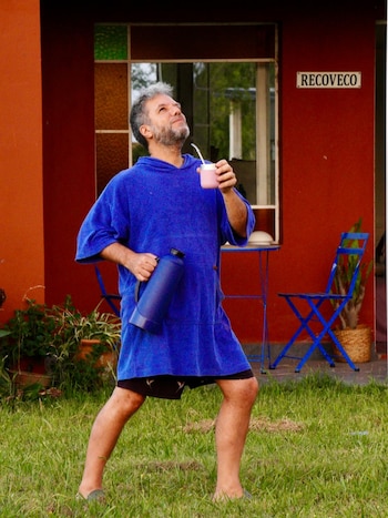 Hombre con barba gris, túnica azul, shorts y sandalias en césped. Sostiene mate rosado y termo azul. Detrás, casa roja, ventana, dos sillas azules, plantas y cartel 'RECOVECO'