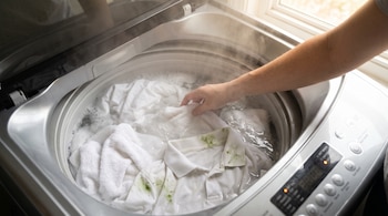 Una mano dentro de una lavadora de carga superior con ropa blanca sucia y manchas de pasto en agua caliente y vapor.