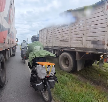 Un motociclista con chaqueta verde y alforjas circula por un espacio estrecho entre dos camiones de carga