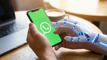 Plano medio de una mano humana sosteniendo un smartphone con el logo de WhatsApp en pantalla, junto a una mano robótica con luces azules, sobre una mesa de madera.