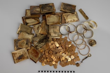 Pila de monedas doradas, varios estuches rectangulares, pulseras metálicas y una regla de medición sobre fondo gris