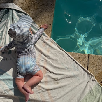 Un bebé con traje de baño de rayas azules y blancas y sombrero a juego yace boca abajo sobre una toalla junto a una piscina