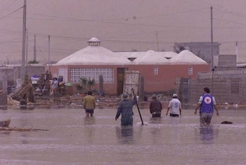 Cinco personas caminan con el agua hasta los muslos en una calle anegada frente a una vivienda y restos de escombros
