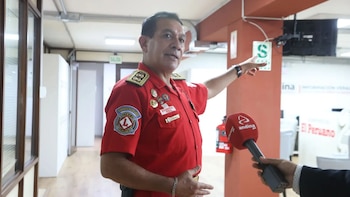 Un hombre con uniforme rojo de bombero con insignias y epaulets apunta mientras es entrevistado con un micrófono rojo de Andina Noticias en un pasillo