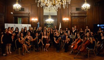 Grupo de mujeres músicas sonrientes posando con violines, violonchelos y clarinetes en una sala lujosa con paneles de madera, arañas de cristal y banderas