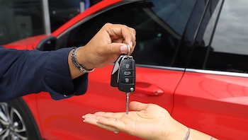 Una mano entrega las llaves de un automóvil sobre la mano abierta de otra persona, frente a la carrocería de un coche rojo