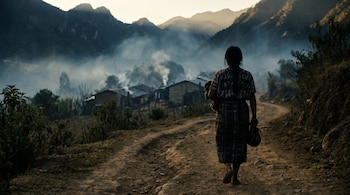 Una persona de espaldas con huipil tradicional camina por un camino de tierra hacia casas rurales en un valle con neblina y montañas.