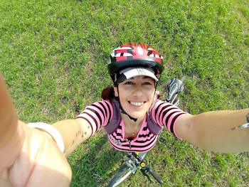 Una mujer con casco de ciclismo y camiseta a rayas rosa y negra se fotografía desde arriba junto a su bicicleta en la hierba