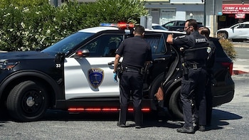 Tres agentes de policía con uniforme, un vehículo policial blanco y negro con luces intermitentes, y edificios comerciales de fondo