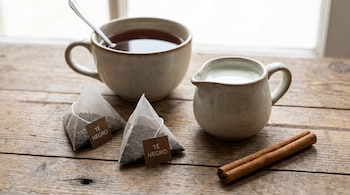 Una taza de té con cuchara, dos saquitos de té negro, una jarra de leche y una rama de canela sobre una mesa de madera rústica junto a una ventana.