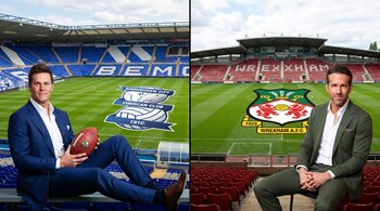 Tom Brady y Ryan Reynolds sentados en trajes, con estadios de fútbol de fondo. Escudos de Birmingham City y Wrexham en el césped.