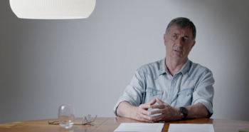 Hombre de mediana edad con camisa vaquera sentado a una mesa de madera, con las manos entrelazadas, frente a una pared gris. Hay un vaso y gafas sobre la mesa