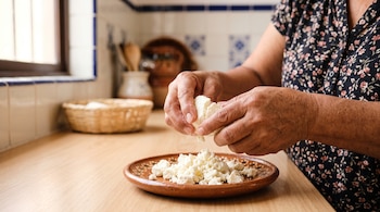 Manos de una mujer desmoronando queso blanco sobre un plato de barro colocado en una mesa de madera.