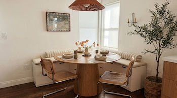 Comedor con mesa redonda de madera, dos sillas con asiento de ratán, un banco acolchado, una lámpara de techo tejida y una planta alta en maceta.