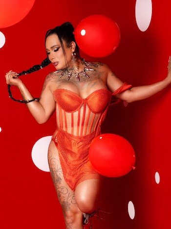 Retrato de Kary Torres de cuerpo completo con atuendo naranja, trenzas largas, collar, tatuajes, sobre fondo rojo con dos globos y círculos blancos