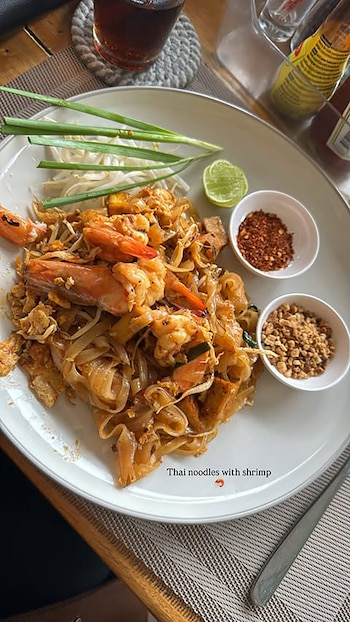 Primer plano de un plato de Pad Thai con camarones, fideos, verduras y tofu, acompañado de lima, cacahuetes triturados y chile en dos pequeños cuencos