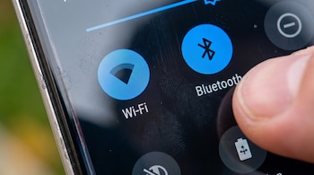 Primer plano de la pantalla de un smartphone con iconos azules de Wi-Fi y Bluetooth y sus nombres. Un dedo se acerca al icono de Bluetooth.