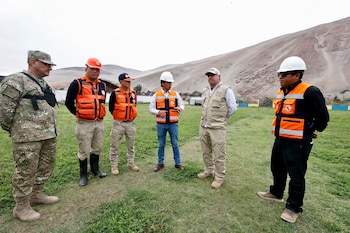 Un militar y cinco funcionarios con chalecos reflectantes y cascos conversan en un campo de hierba frente a cerros áridos