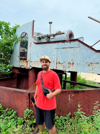 Hombre barbudo con gorra, camiseta roja y bermudas negras de pie ante una estructura metálica oxidada, rodeado de vegetación