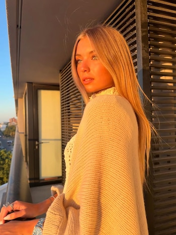 Renata Mónaco, joven rubia, mira de perfil en un balcón. Luz dorada del atardecer ilumina su rostro y cabello. Viste un suéter claro. Al fondo, vista urbana borrosa