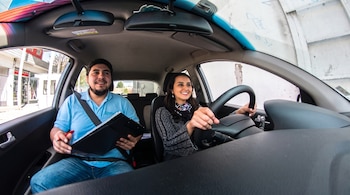 Una mujer sostiene el volante de un coche mientras un hombre en el asiento del copiloto sujeta una carpeta y un bolígrafo.