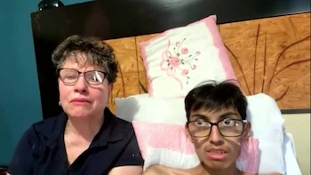 Primer plano de una mujer de mediana edad con gafas y un joven de complexión delgada con gafas, ambos mirando al frente. Hay almohadas detrás