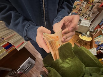 Primer plano de dos manos sosteniendo una pila de billetes de cien pesos argentinos. Al fondo, un sillón verde, un suelo de madera y libros apilados